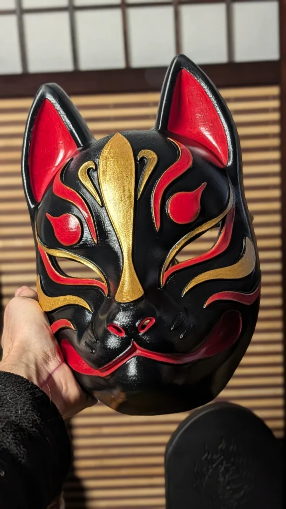 Masque Kitsune noir moderne tenu en main, tracés or et rouge vif, finition vernis brillant, Dai Yokai